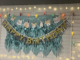 A decorative setup with a 'Happy Birthday' banner featuring glittery gold and silver letters. Above it are cartoon animal faces wearing party hats, and the backdrop is a fringe of metallic teal material. String lights with warm yellow and multicolor fixtures add a festive atmosphere.