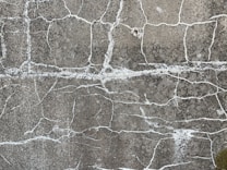 A close-up view of a rough, cracked concrete surface. The surface features intricate patterns of white lines, formed by the cracks spreading throughout the grayish texture.