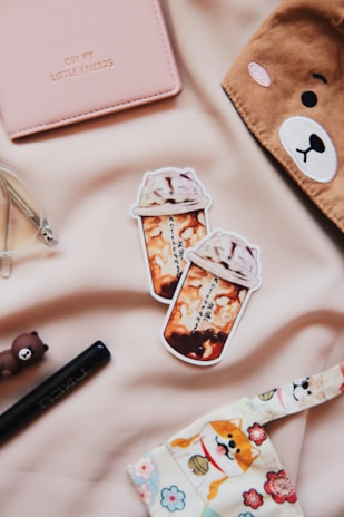 A soft pink notebook with the text 'OH MY LITTLE FRIENDS' embossed on it is placed on a beige surface. Nearby are two stickers depicting cups of bubble tea with foam on top. A brown item with a bear face design adorns the upper right corner, and a small brown bear figurine sits beside a black pen. A decorative fabric with a dog and floral pattern is partially visible at the bottom.