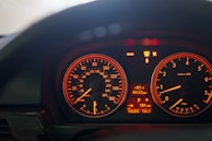 A close-up view of a car dashboard with illuminated speedometer and tachometer gauges. The speedometer shows readings in both miles per hour and kilometers per hour, while the tachometer displays engine speed in revolutions per minute. Various warning and informational lights are visible, such as 'Brake' and 'Service Engine Soon'. The central digital display indicates the temperature, time, and additional vehicle information.