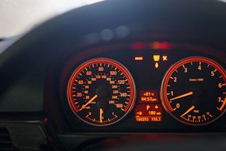A close-up view of a car dashboard with illuminated speedometer and tachometer gauges. The speedometer shows readings in both miles per hour and kilometers per hour, while the tachometer displays engine speed in revolutions per minute. Various warning and informational lights are visible, such as 'Brake' and 'Service Engine Soon'. The central digital display indicates the temperature, time, and additional vehicle information.