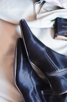 A pair of sleek black leather high-heeled ankle boots with pointed toes and zippers on the side. The boots are placed on a light-colored fabric background, with a small black box positioned nearby.