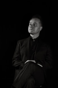 A man in a suit is sitting with a contemplative expression, arms crossed and legs crossed, against a dark background. The lighting highlights his face and upper body, creating a dramatic, monochromatic effect.