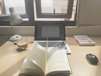 A workspace with a laptop displaying a document, an open book, a lamp, a glass of coffee, and a book titled 'Atomic Habits' on a wooden desk. Sunlight is coming through a window, casting natural light over the setup.