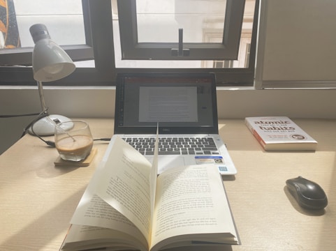A workspace with a laptop displaying a document, an open book, a lamp, a glass of coffee, and a book titled 'Atomic Habits' on a wooden desk. Sunlight is coming through a window, casting natural light over the setup.