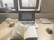 A workspace with a laptop displaying a document, an open book, a lamp, a glass of coffee, and a book titled 'Atomic Habits' on a wooden desk. Sunlight is coming through a window, casting natural light over the setup.