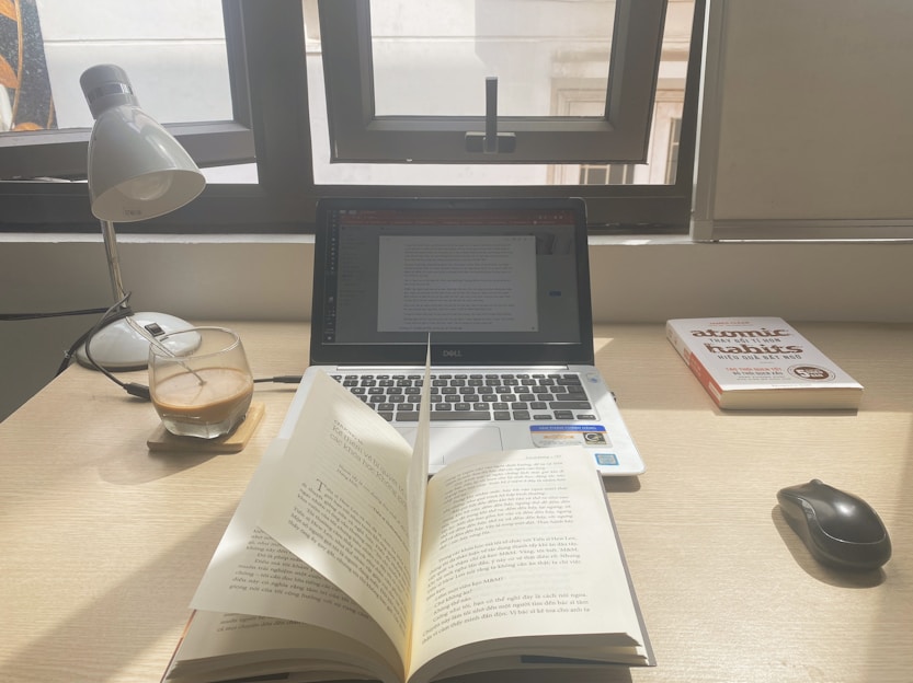 A workspace with a laptop displaying a document, an open book, a lamp, a glass of coffee, and a book titled 'Atomic Habits' on a wooden desk. Sunlight is coming through a window, casting natural light over the setup.