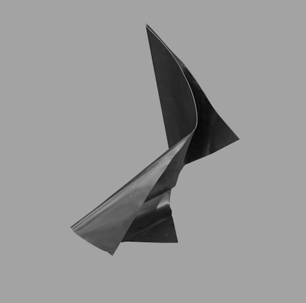 A twisted, abstract metallic sculpture with sharp angles and smooth curves set against a plain gray background. The surface appears smooth and reflects light subtly.