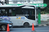 A modern public bus is parked at a bus stop with its side adorned by decorative purple graphics. The bus displays the hashtag '#PilihanCerdasBermobilitas' and is branded with 'BIS-KITA.' In front of the bus, an orange traffic cone is visible. The bus stop is marked by a green and black sign with logos. The background includes a wall with street art and graffiti.