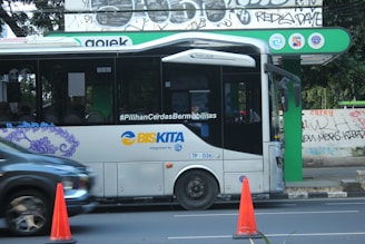 A modern public bus is parked at a bus stop with its side adorned by decorative purple graphics. The bus displays the hashtag '#PilihanCerdasBermobilitas' and is branded with 'BIS-KITA.' In front of the bus, an orange traffic cone is visible. The bus stop is marked by a green and black sign with logos. The background includes a wall with street art and graffiti.
