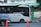 A modern public bus is parked at a bus stop with its side adorned by decorative purple graphics. The bus displays the hashtag '#PilihanCerdasBermobilitas' and is branded with 'BIS-KITA.' In front of the bus, an orange traffic cone is visible. The bus stop is marked by a green and black sign with logos. The background includes a wall with street art and graffiti.