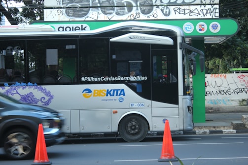 A modern public bus is parked at a bus stop with its side adorned by decorative purple graphics. The bus displays the hashtag '#PilihanCerdasBermobilitas' and is branded with 'BIS-KITA.' In front of the bus, an orange traffic cone is visible. The bus stop is marked by a green and black sign with logos. The background includes a wall with street art and graffiti.