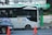 A modern public bus is parked at a bus stop with its side adorned by decorative purple graphics. The bus displays the hashtag '#PilihanCerdasBermobilitas' and is branded with 'BIS-KITA.' In front of the bus, an orange traffic cone is visible. The bus stop is marked by a green and black sign with logos. The background includes a wall with street art and graffiti.