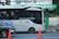 A modern public bus is parked at a bus stop with its side adorned by decorative purple graphics. The bus displays the hashtag '#PilihanCerdasBermobilitas' and is branded with 'BIS-KITA.' In front of the bus, an orange traffic cone is visible. The bus stop is marked by a green and black sign with logos. The background includes a wall with street art and graffiti.