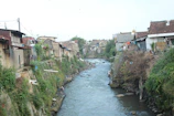 A small river flows through a densely populated area, bordered by various rustic houses. The riverbanks are covered in lush greenery, and some houses are built precariously close to the water. The area looks somewhat overgrown and untidy, with various plants and vegetation climbing the walls. The roofs and walls of the houses are varied in color and material, suggesting a modest community setting.