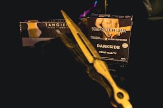 A close-up shot of a dark product box is placed on a reflective surface. The box has text in a foreign language along with English text 'DARKSIDE FRUITTALITY.' A pair of metal tongs or scissors is positioned diagonally across the frame, partially obscuring the box. The background is dark, giving the image a moody and mysterious ambiance.