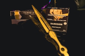 A close-up shot of a dark product box is placed on a reflective surface. The box has text in a foreign language along with English text 'DARKSIDE FRUITTALITY.' A pair of metal tongs or scissors is positioned diagonally across the frame, partially obscuring the box. The background is dark, giving the image a moody and mysterious ambiance.