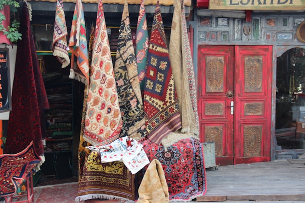 A collection of colorful, patterned rugs and carpets hanging outside a shop with a rustic facade. The shop has a prominently red, ornately carved wooden door. The assortment of textiles includes a variety of traditional designs and motifs, with vibrant reds, blues, and earthy tones. Some greenery can be seen on the left side.