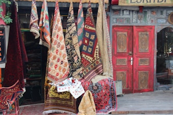 A collection of colorful, patterned rugs and carpets hanging outside a shop with a rustic facade. The shop has a prominently red, ornately carved wooden door. The assortment of textiles includes a variety of traditional designs and motifs, with vibrant reds, blues, and earthy tones. Some greenery can be seen on the left side.
