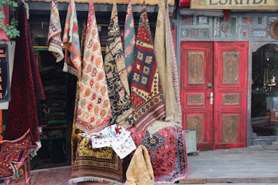A collection of colorful, patterned rugs and carpets hanging outside a shop with a rustic facade. The shop has a prominently red, ornately carved wooden door. The assortment of textiles includes a variety of traditional designs and motifs, with vibrant reds, blues, and earthy tones. Some greenery can be seen on the left side.