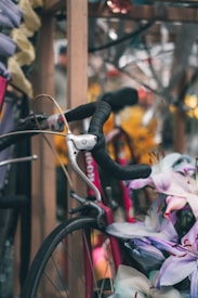 A bicycle handlebar is partially visible with a black grip and silver brake lever. Surrounding it, there are light purple and pink flowers arranged closely together, contrasting with the muted colors of the background and the vibrant pink of the bicycle frame.