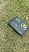 A solar charge controller is placed on grass, featuring a green and black design with various icons and text indicating its functionality. The device includes symbols for solar panels, battery, and other power-related functions.