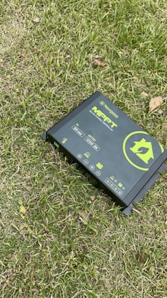 A solar charge controller is placed on grass, featuring a green and black design with various icons and text indicating its functionality. The device includes symbols for solar panels, battery, and other power-related functions.