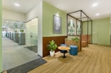 A modern office space features a welcoming waiting area with wooden floors. The area includes a small round table surrounded by two blue ottomans and greenery in planters. Artistic wall decorations with botanical patterns complement the soft green walls. In the background, a glass door leads to an open-plan office with workstations and additional plants, creating a fresh and inviting atmosphere.