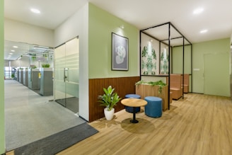 A modern office space features a welcoming waiting area with wooden floors. The area includes a small round table surrounded by two blue ottomans and greenery in planters. Artistic wall decorations with botanical patterns complement the soft green walls. In the background, a glass door leads to an open-plan office with workstations and additional plants, creating a fresh and inviting atmosphere.