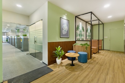 A modern office space features a welcoming waiting area with wooden floors. The area includes a small round table surrounded by two blue ottomans and greenery in planters. Artistic wall decorations with botanical patterns complement the soft green walls. In the background, a glass door leads to an open-plan office with workstations and additional plants, creating a fresh and inviting atmosphere.