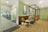A modern office space features a welcoming waiting area with wooden floors. The area includes a small round table surrounded by two blue ottomans and greenery in planters. Artistic wall decorations with botanical patterns complement the soft green walls. In the background, a glass door leads to an open-plan office with workstations and additional plants, creating a fresh and inviting atmosphere.