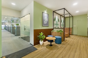 A modern office space features a welcoming waiting area with wooden floors. The area includes a small round table surrounded by two blue ottomans and greenery in planters. Artistic wall decorations with botanical patterns complement the soft green walls. In the background, a glass door leads to an open-plan office with workstations and additional plants, creating a fresh and inviting atmosphere.