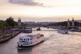 A river scene with a large white tour boat named 'RIVER'S KING' navigating along the water. In the background, an ornate bridge is adorned with golden statues, leading towards a city skyline. The sky is a soft gradient of sunset hues with scattered clouds, creating a calm evening atmosphere. Both sides of the river are lined with people walking and lush trees.