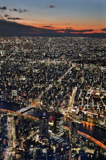 A sprawling cityscape illuminated by countless city lights as dusk sets in, with a vibrant sky blending hues of orange, red, and blue. A river runs through the city, crossed by several lit bridges, and numerous skyscrapers and buildings are visible.