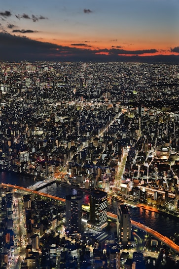 A sprawling cityscape illuminated by countless city lights as dusk sets in, with a vibrant sky blending hues of orange, red, and blue. A river runs through the city, crossed by several lit bridges, and numerous skyscrapers and buildings are visible.