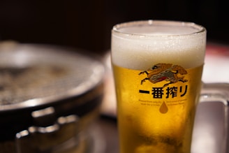 A frothy glass of beer in a clear mug with an illustrated logo featuring a stylized creature and Japanese characters. The background includes a blurred view of a round grill, suggesting a dining or bar setting.