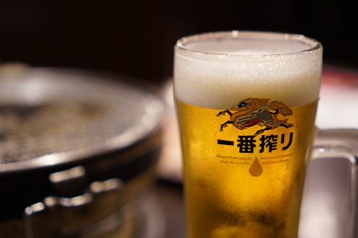 A frothy glass of beer in a clear mug with an illustrated logo featuring a stylized creature and Japanese characters. The background includes a blurred view of a round grill, suggesting a dining or bar setting.