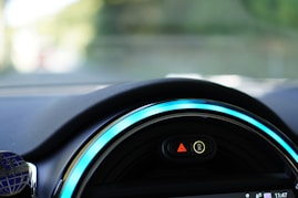 Close-up view of a car dashboard featuring a digital display with illuminated symbols. The dashboard is sleek with vibrant blue accent lighting encircling the display. The symbols include a hazard warning triangle and an indicator for unfastened seatbelts.