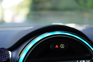 Close-up view of a car dashboard featuring a digital display with illuminated symbols. The dashboard is sleek with vibrant blue accent lighting encircling the display. The symbols include a hazard warning triangle and an indicator for unfastened seatbelts.