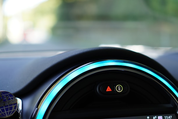 Close-up view of a car dashboard featuring a digital display with illuminated symbols. The dashboard is sleek with vibrant blue accent lighting encircling the display. The symbols include a hazard warning triangle and an indicator for unfastened seatbelts.