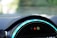 Close-up view of a car dashboard featuring a digital display with illuminated symbols. The dashboard is sleek with vibrant blue accent lighting encircling the display. The symbols include a hazard warning triangle and an indicator for unfastened seatbelts.