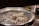 A close-up of a metal grill with glowing charcoal, possibly part of a barbecue or cooking setup. In the background, there are blurred items that appear to be bowls with food or garnishes.