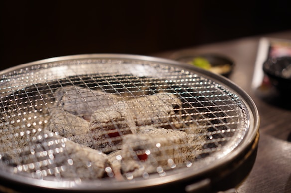 A close-up of a metal grill with glowing charcoal, possibly part of a barbecue or cooking setup. In the background, there are blurred items that appear to be bowls with food or garnishes.