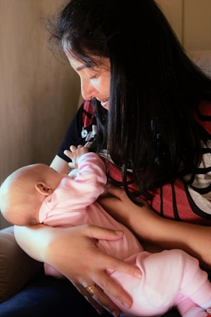 A woman is holding a baby in her lap in a nurturing manner. The woman has long dark hair and is wearing a red and black top with a ring on her finger. The baby is dressed in a pink outfit and appears to be breastfeeding.