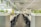 A modern office environment with a long row of workstations featuring sleek black chairs and monitors. Lush green plants are arranged on partitions and overhead, adding a refreshing touch to the otherwise minimalistic design. The ceiling lights provide sufficient illumination, highlighting the clean and organized workspace.