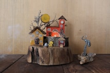 A miniature village crafted on a wooden base featuring several small houses, each uniquely designed with elements like clocks, windows, and roofs. A prominent red tower stands in the background adorned with a clock face. Bare, wire-like tree branches protrude from the base along with other small decorative objects, including a figure resembling a stickman made from wires and a small piece of art in the form of abstract objects. The atmosphere appears whimsical and artistic, with a blend of rustic and fantastical themes.