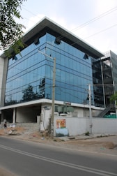 A modern glass office building with a reflective facade, set against a backdrop of clear sky. The structure has multiple floors with large windows, and there is a construction area in front with debris. An electrical pole is situated near the building, and a poster is attached to a low white wall.
