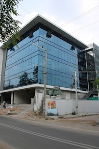 A modern glass office building with a reflective facade, set against a backdrop of clear sky. The structure has multiple floors with large windows, and there is a construction area in front with debris. An electrical pole is situated near the building, and a poster is attached to a low white wall.
