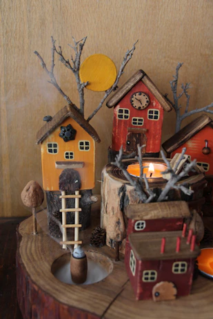 A whimsical miniature scene featuring small wooden houses with painted details, rustic chimneys, and tree branches. Yellow and red houses are arranged on a circular wooden base, surrounded by small twigs and a candle in the center emitting a warm glow. A tiny ladder leads to the entrance of the orange house.