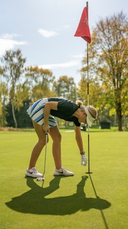 A golfer is bending down on a green putting surface, reaching for a golf ball near the hole. The golfer is wearing a visor, shorts, and golf gloves, holding a putter in the other hand. A red flagstick stands upright in the hole, surrounded by lush green grass and trees with autumn foliage in the background.
